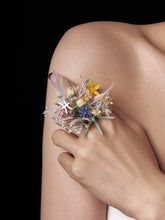 A hand adorned with a Pisces Flower Ring by CAMELLIA, crafted in New York, gently holds a bouquet of small, colorful flowers and delicate stems against bare skin.